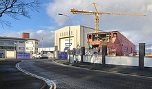 Picture of Red ship hull under construction with a building to the left of the image and a metal fence in front