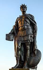 A close up photograph of a sculpture of a standing crowned man, his left hand is on his sword hilt while his shield is at his left leg