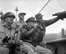 MacArthur is seated, wearing his field marshal's hat and a bomber jacket, and holding a pair of binoculars. Four other men also carrying binoculars stand behind him.