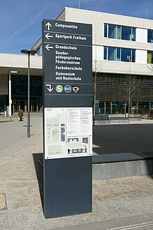 Schwarze Infosäule mit Beschriftungen und Pfeilen in weiß zur Campusmitte, dem Sportpark Freiham und verschiedenen Schulen, Richtungspfeil mit S-Bahn und Bussymbol und darunter einem Lageplan des Campus. Hinter der Säule sieht man moderne Gebäude.