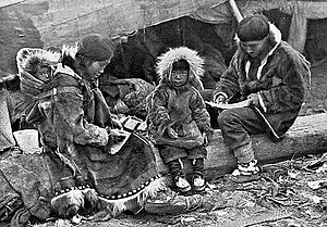 An Inuit family is sitting on a log outside their tent. The parents, wearing warm clothing made of animal skins, are engaged in domestic tasks. Between them sits a toddler, also in skin clothes, staring at the câmera. On the mother's back is a baby in a papoose.