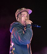 A man in a black cap and hoodie with a microphone in his hand rapping on stage