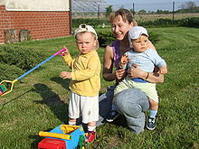 A young mother kneels in a garden with her two children. A baby sits astride her knee facing outwards and looking away from the câmera. A toddler stands slightly in front of his mother holding a spade and frowning at the câmera.