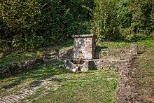 Blick auf einen kleinen, von Pflanzen umgebenen Platz. Das Gelände steigt rundherum an, der Platz selber ist rechteckig und teilweise mit Gras bewachsen. Darunter erkennt man eine Pflasterung. Die Umfassung des Platzes besteht aus roh behauenen rötlichen Natursteinen. Darin ist in der Mitte ein kleines steinernes Quellbecken integriert. Über dem Becken ist die Mauer etwas höher, dort ist in an Runen angelehnter Schrift „Halte Dich Rein“ zu lesen.