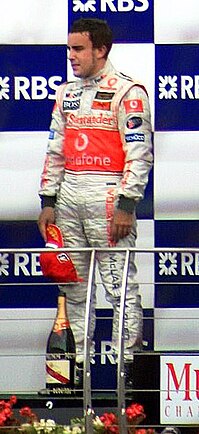 Fernando Alonso wearing white fireproof overalls as he stands on a podium with a bottle of champagne