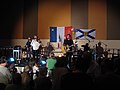 Photographie du groupe de musique Suroît durant l'un de ses spectacles. Sur le mur au fond de la scène se trouvent le drapeau de l'Acadie et celui de la Nouvelle-Écosse.