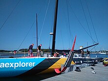 Vu de trois quarts avant, un voilier noir, bleu et jaune amarré au ponton. Bout-dehors et foils rouges. Mât noir.