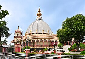 A tall ornate domed structure, topped with a spear and flanked by green lush trees.