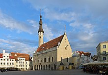 Seitliche Farbfotografie einer hellbraunen Kirche mit schmaler Turmlaterne und rotem Dach. Das Erdgeschoss ist mit mehreren Bögen durchzogen, über denen schmale Fenstern liegen. Der Turm hat eine lange, dunkle Spitze. Um das Gebäude sind Häuser und gastronomische Außenterrassen. Der Platz ist im linken Vordergrund leer.