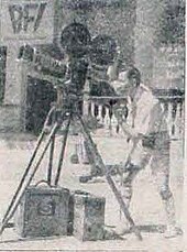 A man with short hair and a white shirt is controlling a large camera. He is facing left.