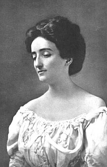 A white woman with her dark hair in an updo, wearing a dress with a low neckline that exposes her shoulders. She is looking downward.