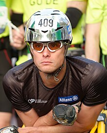 Headshot of a white man with black top, sunglasses and a silver helmet.