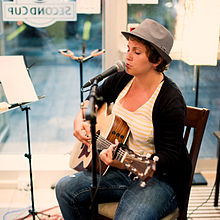 A photograph of a person wearing a grey hat, a yellow-and-white striped shirt, and blue jeans playing a guitar and singing into a microphone