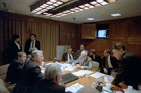 Des hommes en costume assis autour d'une table ovale discutent. Des papiers sont sur la table. Au fond, une télévision des années 1980 est allumée.