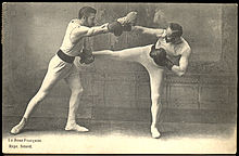 Photographie en noir et blanc de deux hommes combattant en intérieur avec des mouvements de boxe française.