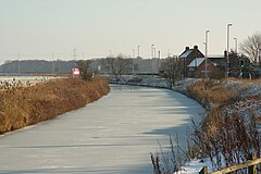 The Fosdyke curves through the picture, its surface smooth white ice. The two banks are brown with dead grass, and the fields either side crop-less and white with hoar. Farther along the bank, in the middle distance, a small group of redbrick buildings are grouped together. Lamp posts are ranged on the right bank, facing away from the canal. They are for lighting the adjacent, but invisible, A57 road.