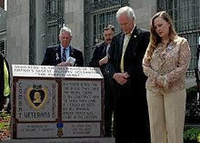 Uma foto colorida de Daniel e Maureen Murphy ao lado de um monumento em frente à Agência Postal dos Estados Unidos Tenente Michael P. Murphy em Patchogue, Nova Iorque. O monumento tem uma Medalha Purple Heart e algumas palavras gravadas nele e há dois homens ao fundo.