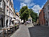 Farbfotografie einer Pflastersteinstraße mit Außentischen auf der linken Seite und alten Häusern an beiden Seiten. In der Mitte wird die Straße von einem Steinmonument und Bäumen aufgeteilt.