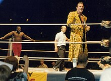 Junkyard Dog se tient debout dans le coin gauche du ring. Au premier plan, le catcheur Brutus Beefcake regarde le public.