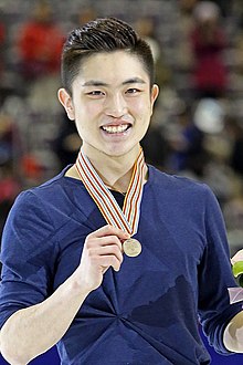 Yan Han trägt ein blaues Oberteil, mit der rechten Hand präsentiert er seine Bronzemedaille, die an einem gestreiften Band um seinen Hals hängt.