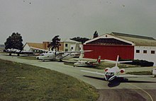 Photographie ancienne représentant quatre avions devant un hangar à la porte rouge. De gauche à droite : un B-17, un Hurel, un AirCommander et un Mystère 20.