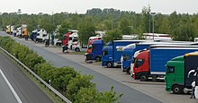 Mehrere Lkws stehen nebeneinander auf einer Betonfläche, dazwischen und dahinter stehen Bäume. Durch eine Leitplanke und eine Reihe Büsche ist die Fläche mit den Lkws von der Autobahn abgetrennt.