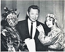 Black and white photograph of Johnny Kan, a restaurateur in Chinatown, San Francisco. Kan is wearing a dark suit with a dark tie. He stands between two actors, a devil on the left side and an angel on the right side, who belong to the Foo Hsing Opera Company.