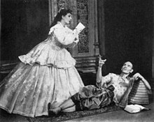 A man with a shaven head, wearing Asian dress, reclines on the floor and gestures at a woman in 19th century dress, who is writing, apparently at the man's dictation.