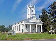 A white church with a colonnaded front and square steeple topped with spiky finials on the corners on a mowed grassy area under a blue sky. Behind it on the right are tall evergreen trees. In front on the left is a sign saying "Reformed Church of New Hurley" and a green street sign with "New Hurley Road" on it