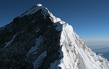 Photographie couleur d'une crête de montagne dont une face, abrupte, est recouverte de neige, et dont l'autre, rocheuse, est escaladée par quelques grimpeurs paraissant minuscules.