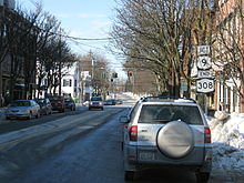 Ground-level view of a road within a small village. The road is lined with moderately sized trees. Snow and ice is present.