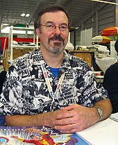 A middle-aged man with graying brown hair, a beard, glasses, and a Three Stooges shirt sitting at a table.