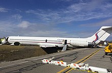 A jet airliner with tail-mounted engines and a T-tailed configuration, painted mostly white with a thin red stripe running along the fuselage below the windows, lying on its belly across a ditch; the aircraft's tail is resting on a paved road, while the forwards portion of the fuselage rests on a grassy field on the other side of the ditch. The aircraft's tailcone has detached and is lying on the grass next to the left side of the fuselage.