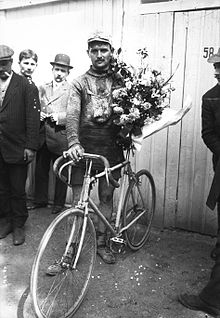 Photographie noir et blanc d'un cycliste debout sur son vélo, portant un bouquet.