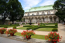 Un bâtiment clair, au toit de cuivre vert, une grande arcade courant face à un jardin fait de pelouse, de buis et de fleurs rouges en pot au premier plan.