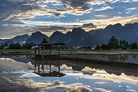 Réflexion dans l'eau d'une hutte en bois avec un ciel coloré de nuages orange, gris et blancs, dans l'eau d'une rizière, avec des montagnes karstiques au coucher du soleil pendant la mousson, dans la compagne de Vang Vieng.