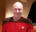 A photograph of a smiling man with white hair who is looking at the viewer, standing in a room, and wearing a red shirt with black shoulders