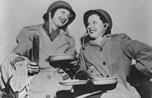 Two woman leaning toward each other are laughing and dressed in World War II oversized military coats and helmets