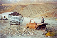 Paysage de montagne quasi désertique. Au premier plan, la carcasse incomplète et rouillée d'un véhicule, vestige de la guerre civile. Au loin, un hangar apparemment désaffecté