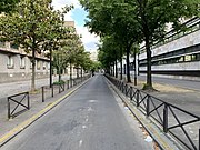 Photo d'une rue bordée d'immeubles de chaque côté, la chaussée est goudronnée et bordée de chaque côté par des barrières et des arbres.