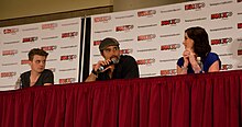 Photograph of three people sitting at a table, one of whom is holding a microphone.