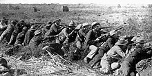 Boer War scene. Men of all ages wearing hats and bandoleers crouch in a line, rifles pointed