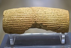 Rear view of a barrel-shaped clay cylinder resting on a stand. The cylinder is covered with lines of cuneiform text