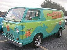 Das Farbfoto zeigt einen Kleintransporter („Van“) in Frontlenker-Bauweise, der in psychedelischen Farben und Mustern im Hippie-Stil lackiert ist. Großflächig dominieren hellblau und hellgrün; Blumenmuster und der Schriftzug „The Mystery Machine“ sind in oranger Farbe aufgebracht. Das Fahrzeug ähnelt sowohl bezüglich der Bauart als auch der Lackierung dem Van der Mystery Inc. aus der ursprünglichen Scooby-Doo-Zeichentrickserie.