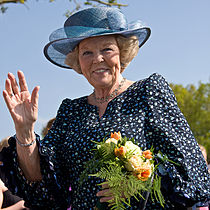 Dronning Beatrix iført hat og blomsterudsmykket kjole, bærer blomster og vinker