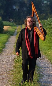 A man wearing black clothing and a red scarf, walking along a gravel path and carrying a harp over his shoulder