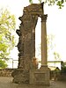 Column and fragment of an arch with an inscription, standing on a cobbled surface with a low wall and fence behind