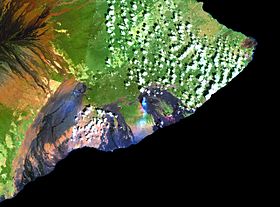 Image satellite du Kīlauea en 2001 mettant en évidence la végétation (en vert et brun), les coulées de lave (en noir) et les panaches volcaniques (nuages bleuâtres) du Halemaʻumaʻu (à gauche) et du Puʻu ʻŌʻō (à droite) ; des coulées de lave du Mauna Loa sont visibles en haut à gauche.