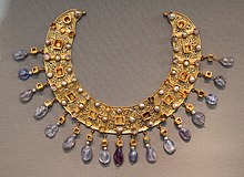 A close up photograph of a piece of golden necklace like jewellery, with multiple beads of varying shades of blue hanging from the central gold plate in a semicircular pattern