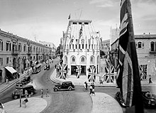 A period photo of street scene. Cars and pedestrians move through an intersection decorated with Union Jacks and Allied flags